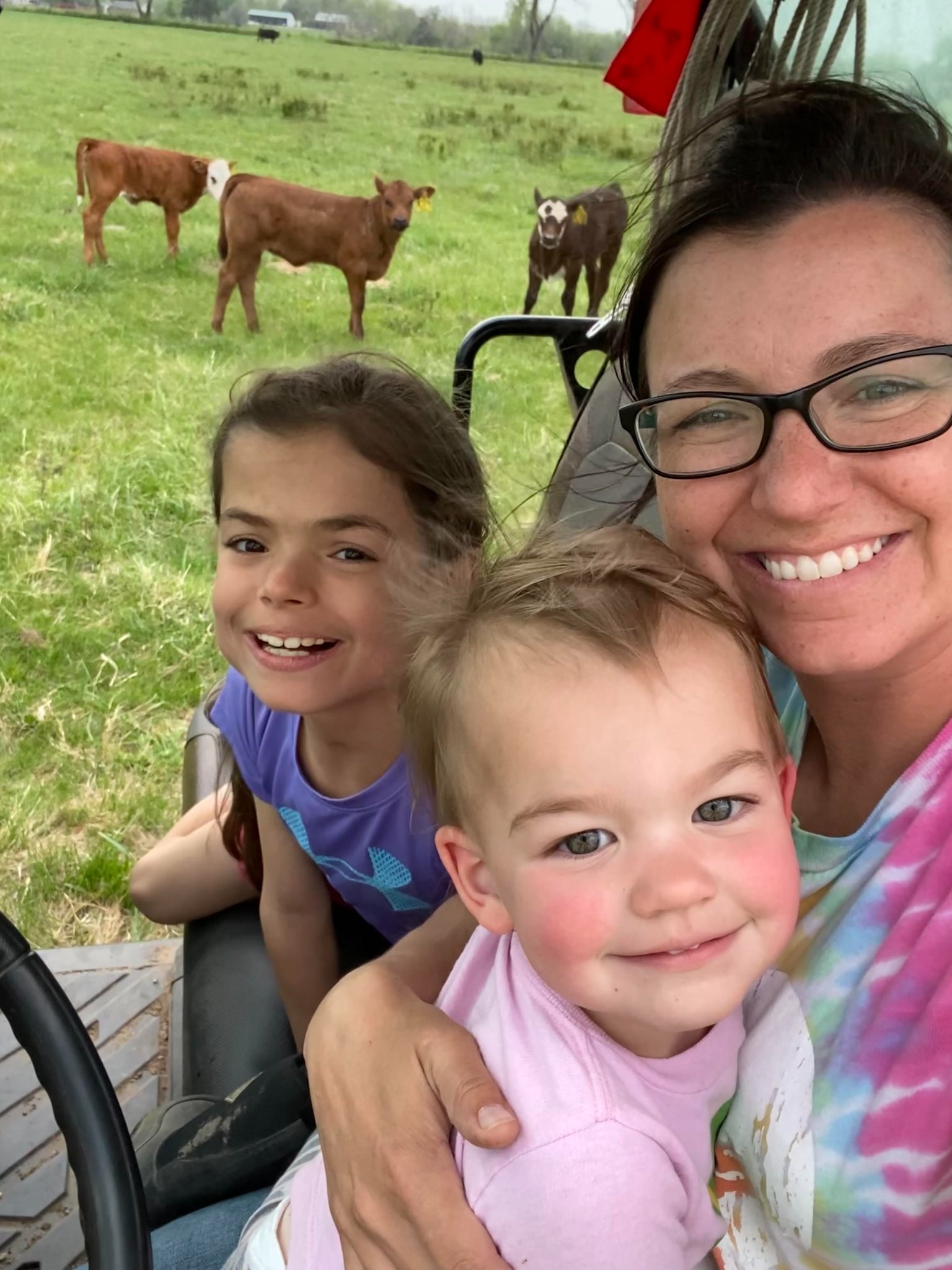 285060931_102179519190953_400429673643237369_n A smiling mother poses with her two young daughters in a tractor on a farm, with calves grazing in the background. The scene captures a joyful family moment in a rural setting, highlighting the connection to agriculture and animal care.