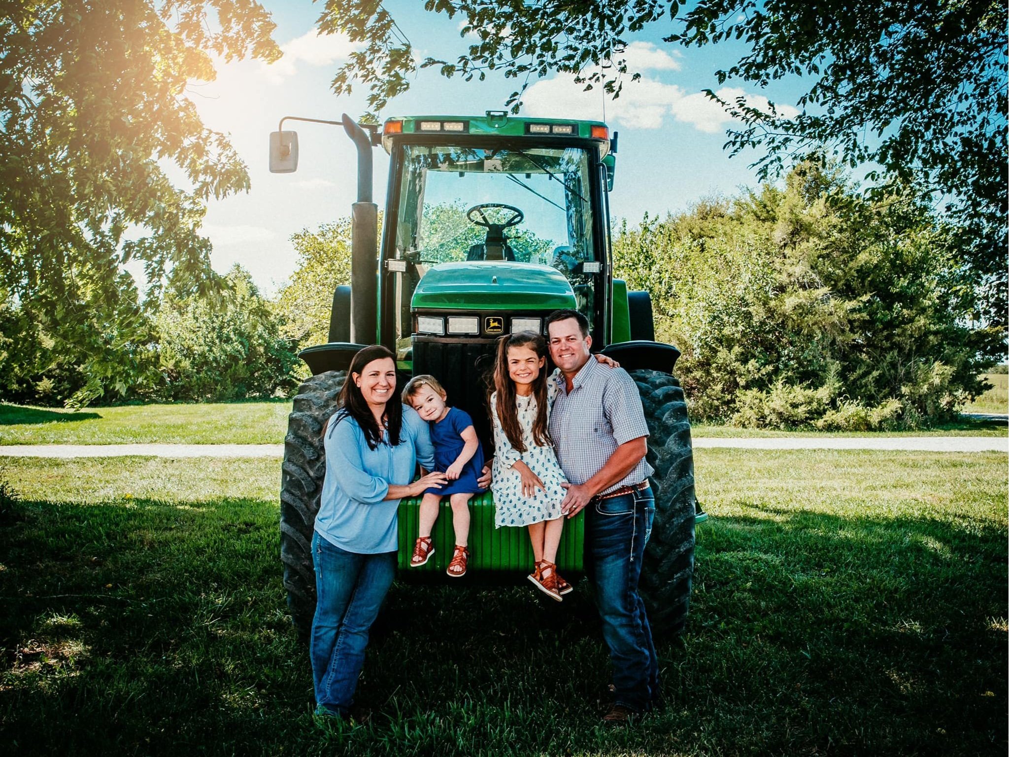 Family portrait in front of a green tractor, featuring a mother, father, and two young daughters, set in a sunny outdoor environment with trees and grass.