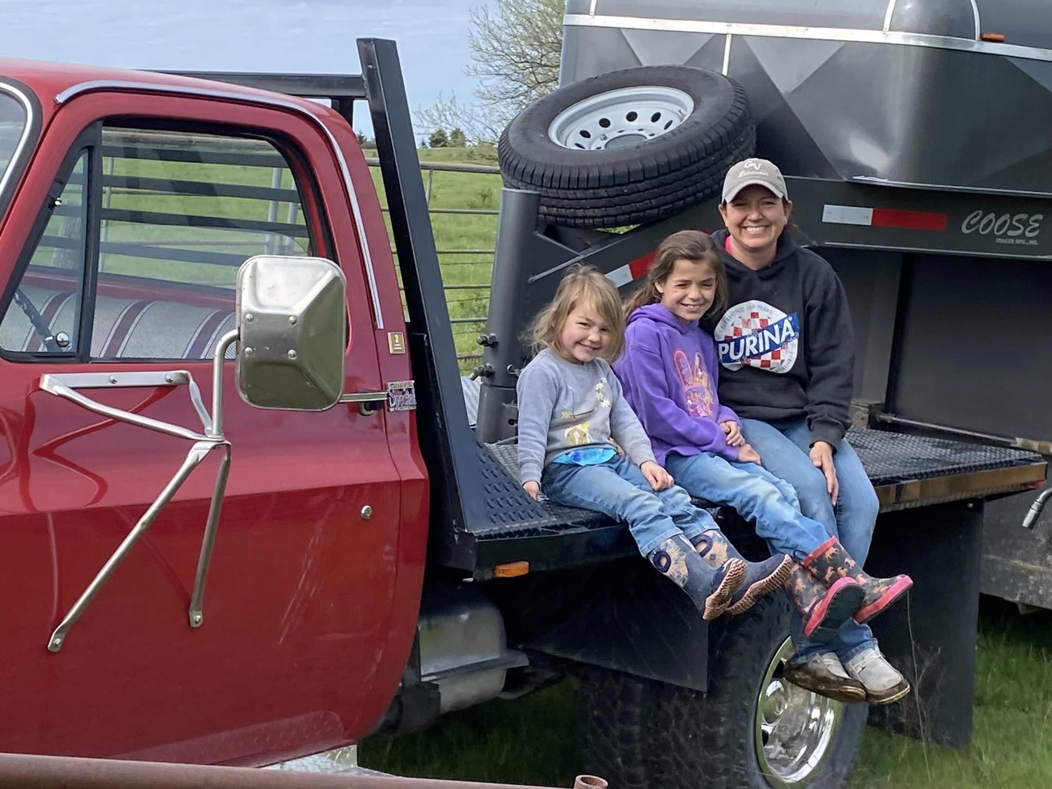 443712617_10230169909810322_1963858726875128667_n A woman and two young girls sitting on the tailgate of a red pickup truck, smiling and enjoying a sunny day outdoors. The truck is parked next to a livestock trailer, with green fields in the background. The girls are wearing colorful boots and casual clothing, while the woman is dressed in a sweatshirt and cap.