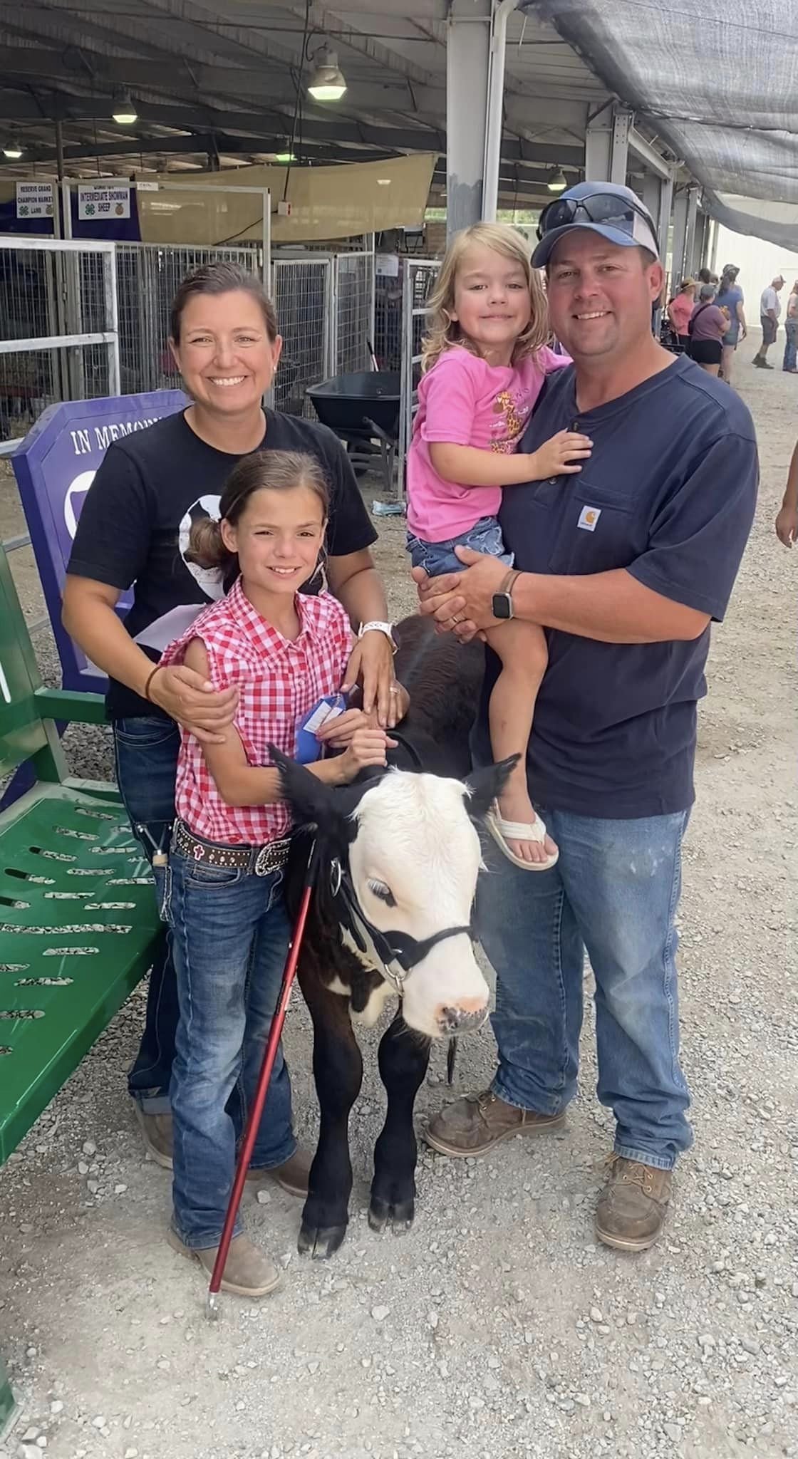 453477898_10230784225687835_1733312493247865723_n Family posing with a calf at a livestock event, showcasing a smiling mother, father, and two young daughters. The scene includes a green bench and livestock pens in the background, emphasizing a family-friendly atmosphere at the fair.