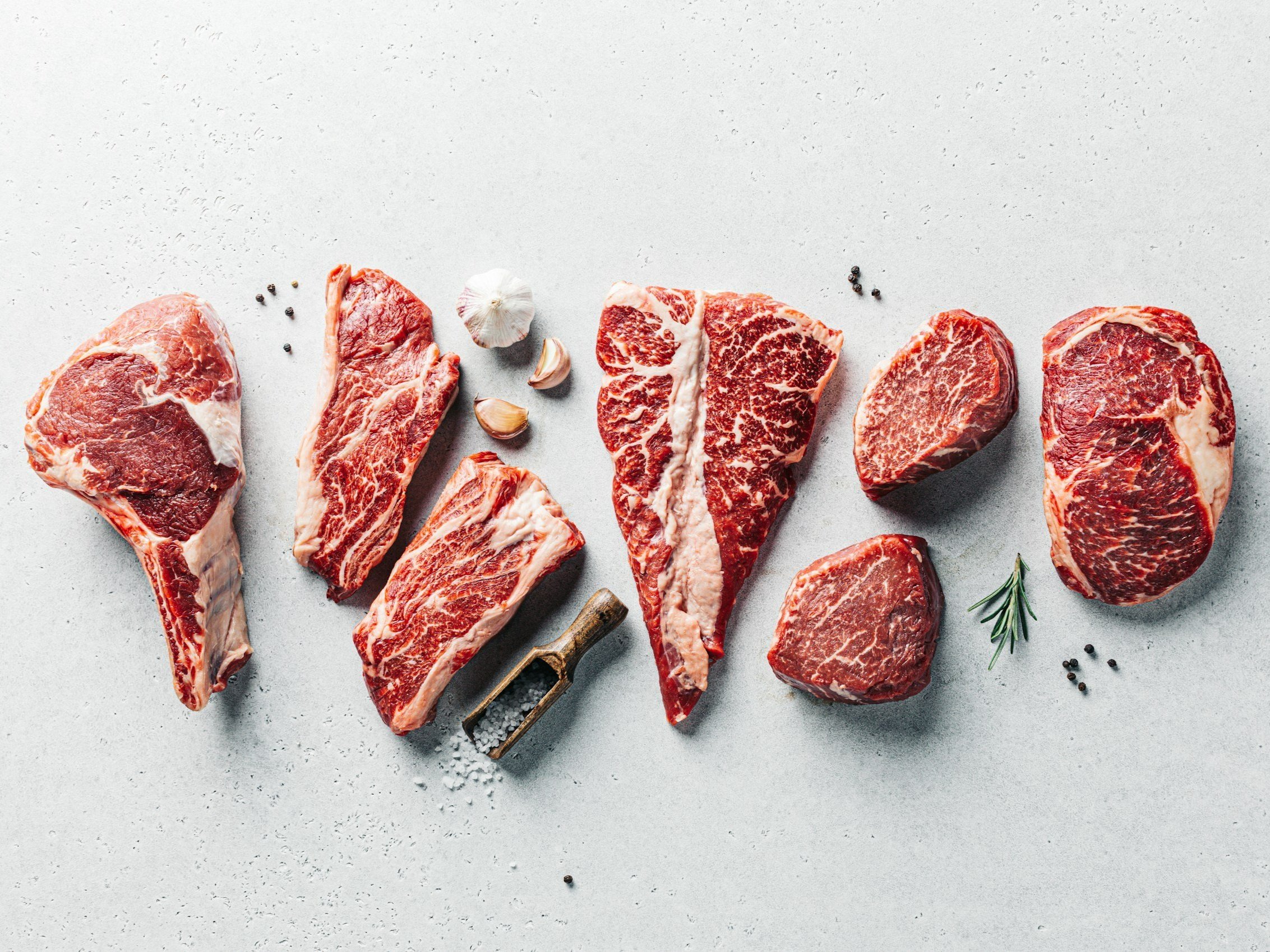 Assorted cuts of raw marbled beef including ribeye, sirloin, and tenderloin, arranged on a light gray surface with garlic cloves, rosemary, and black peppercorns for seasoning.