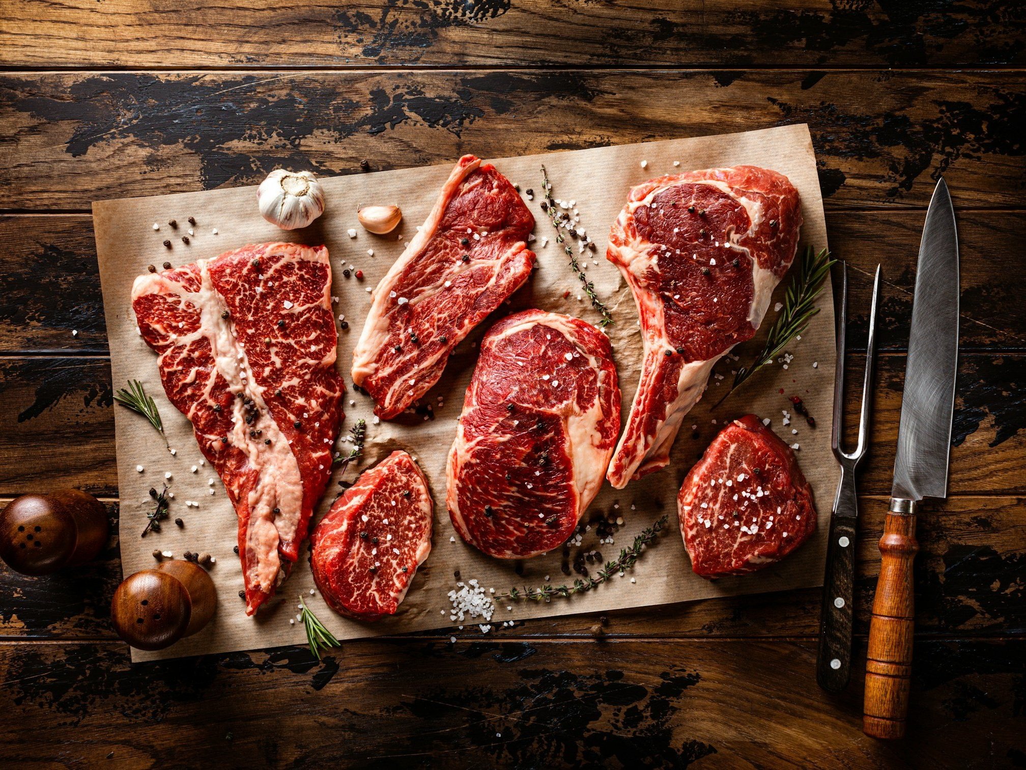 Assorted cuts of marbled beef on parchment paper, garnished with herbs, garlic, and seasoning, with knives and salt shakers on a rustic wooden table.